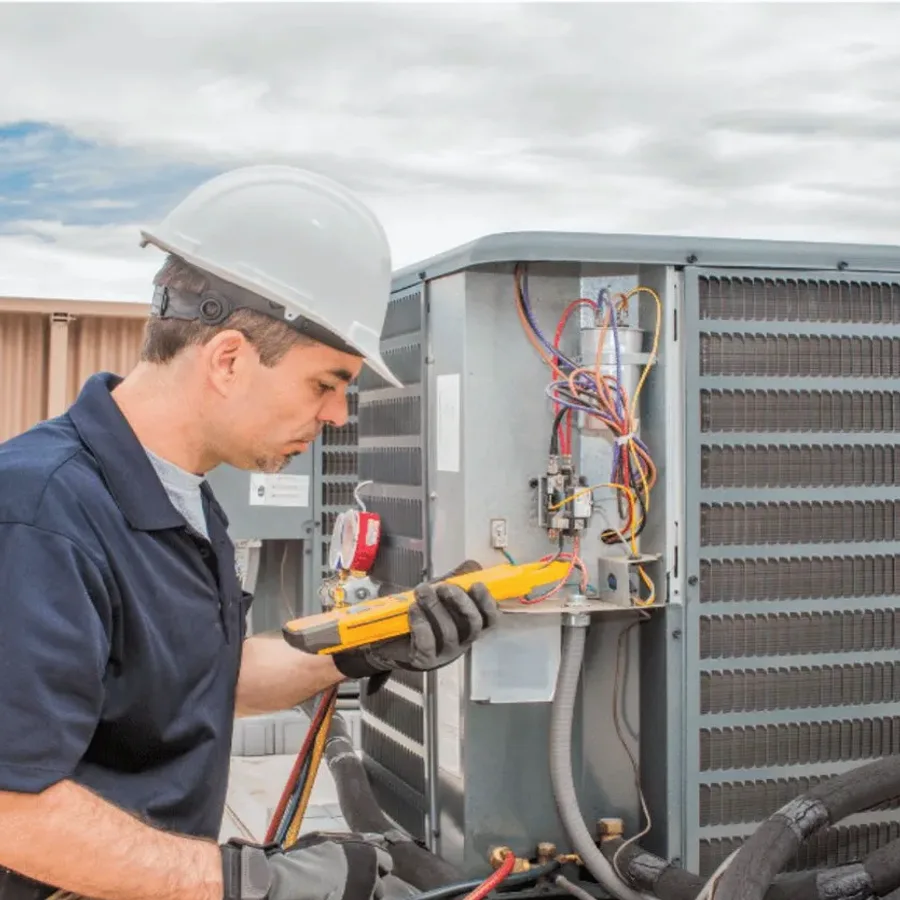 HVAC technician in a hard hat testing an outdoor air conditioner with a multimeter